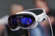 Headset de VR/AR branco e preto em um suporte. A frente espelhada reflete pessoas e luz azul.