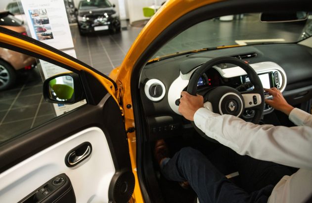 Pessoa em carro amarelo com porta aberta, mãos no volante e painel, em ambiente de concessionária.