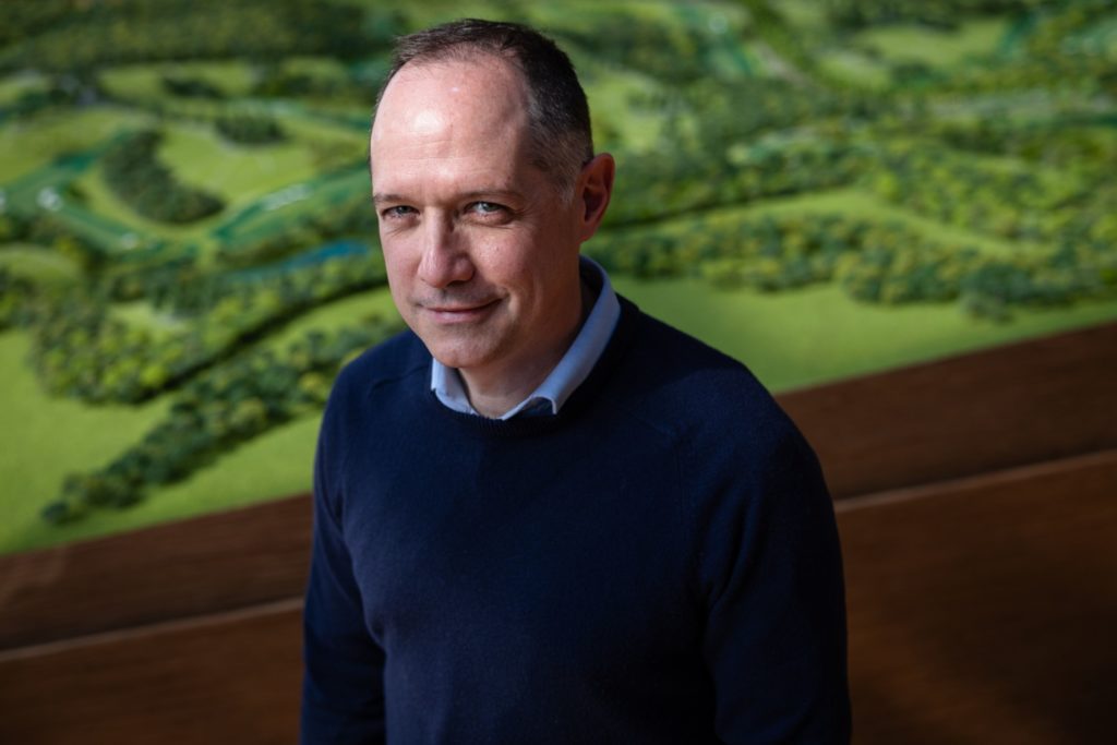 Homem sorridente em suéter azul, com modelo de paisagem verde ao fundo.