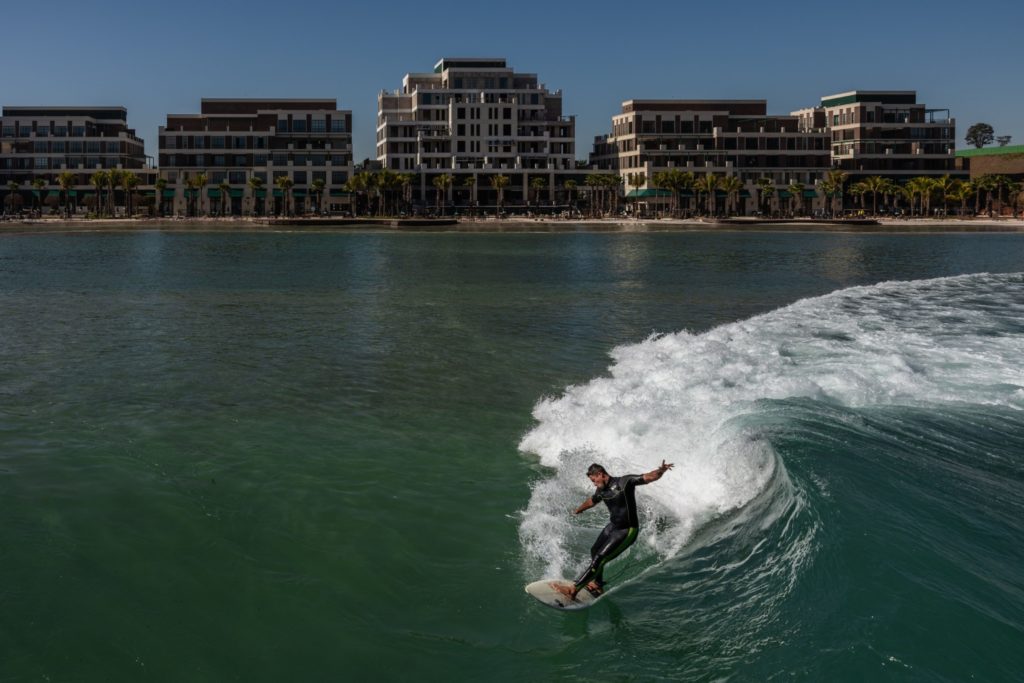 Surfista em onda gerada, com edifícios e palmeiras ao fundo.