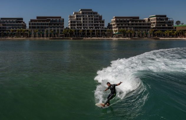 Surfista em onda gerada, com edifícios e palmeiras ao fundo.