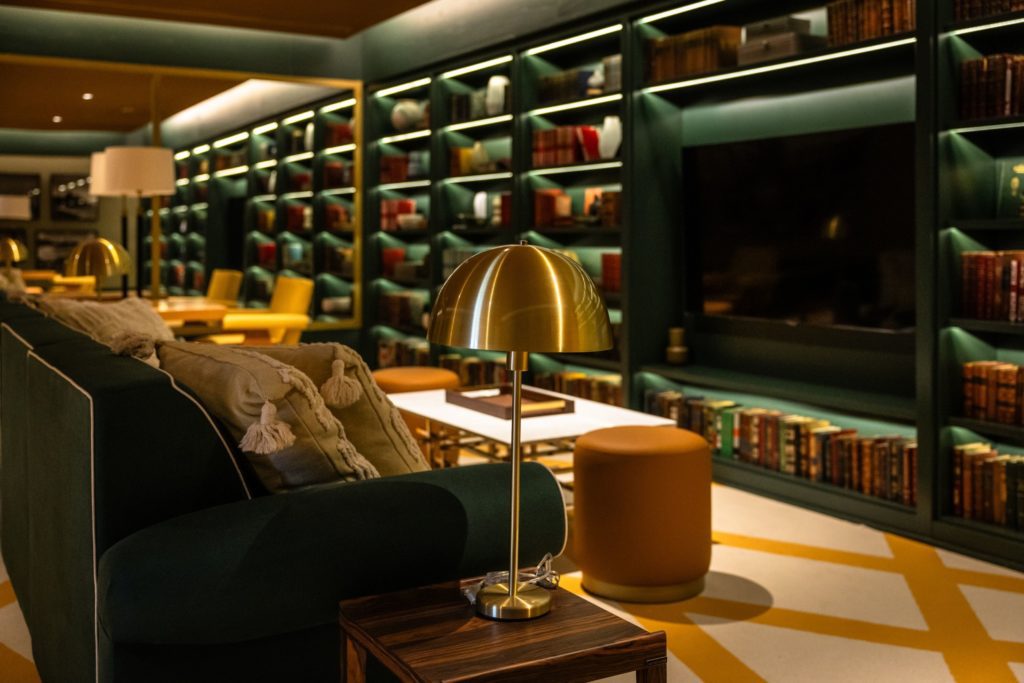 Lounge moderno com estante de livros verde, sofá, luminária dourada e piso geométrico.