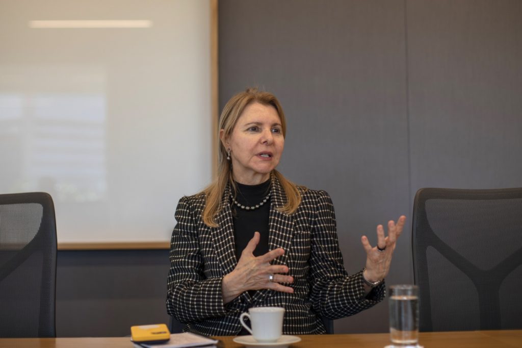 Mulher loira de blazer xadrez gesticula ao falar em reunião.