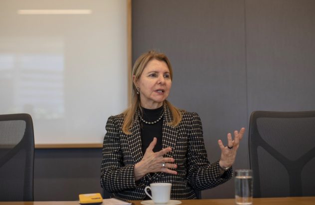 Mulher loira de blazer xadrez gesticula ao falar em reunião.