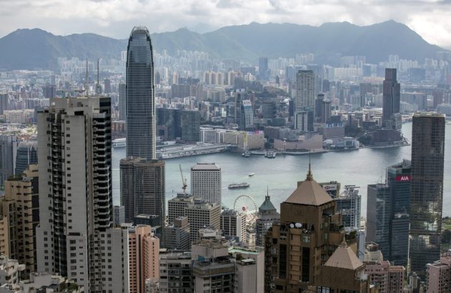 Hong Kong: Densa paisagem urbana com arranha-céus altos, barcos no porto e montanhas ao fundo sob um céu nublado.