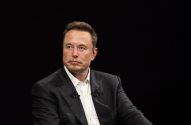 Elon Musk de terno escuro e camisa branca, sentado com olhar pensativo.
