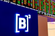 Logo da B3 em tela azul, com painel de cotações da bolsa de valores exibindo dados financeiros ao fundo.