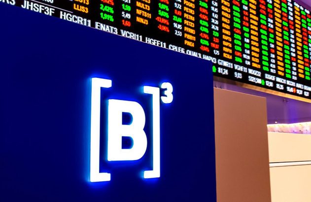 Logo da B3 em tela azul, com painel de cotações da bolsa de valores exibindo dados financeiros ao fundo.