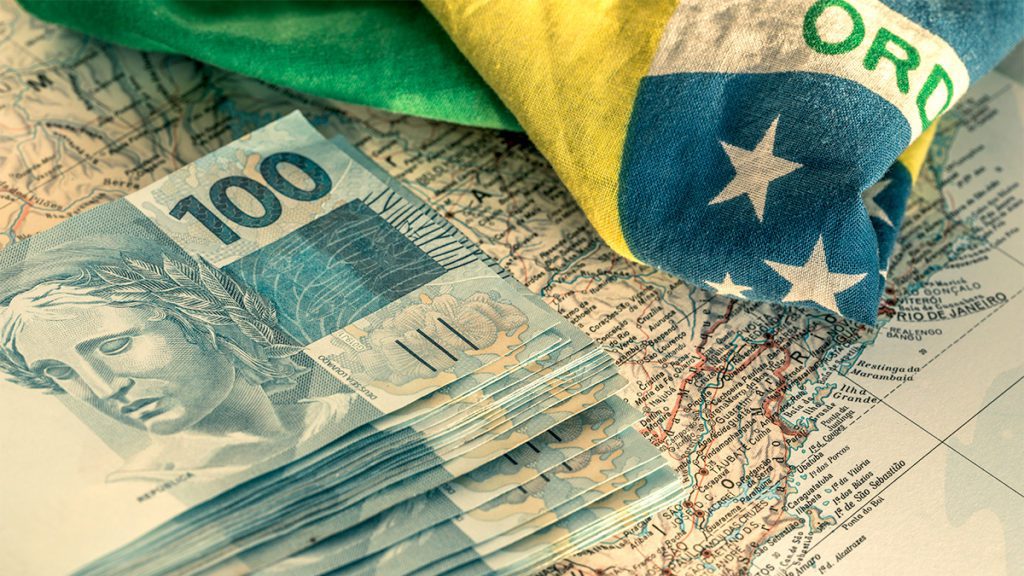 Notas de 100 reais e bandeira do Brasil sobre um mapa.