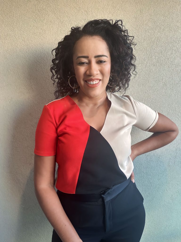 Mulher sorrindo, cabelo cacheado, em pé, vestindo blusa color-block e calça escura, mão na cintura.