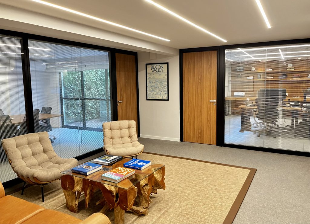 Escritório moderno com área de estar, poltronas, mesa, e salas de trabalho com divisórias de vidro.