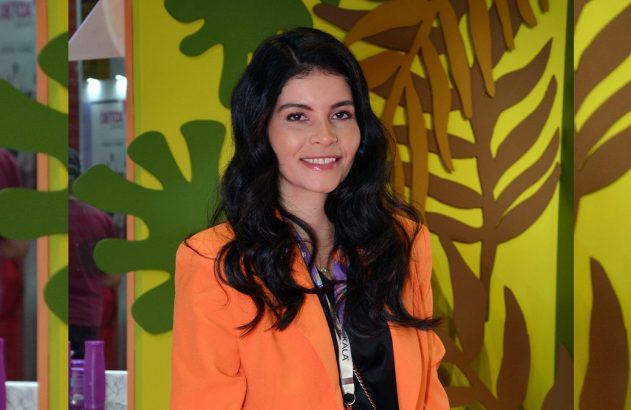 Mulher sorrindo com longos cabelos escuros e blazer laranja. Fundo vibrante com recortes de folhagens.