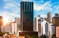 Paisagem urbana ensolarada: arranha-céus e edifícios sob céu azul com nuvens. Brilho forte do sol à esquerda.