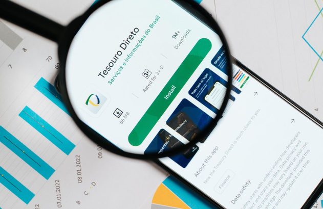Lupa sobre tela de celular exibindo página de aplicativo Tesouro Direto, com gráficos financeiros ao fundo.