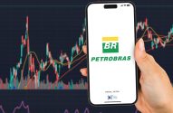 Mão segura celular com logo Petrobras (PETR3/PETR4) na tela. Ao fundo, gráfico de ações da bolsa de valores.