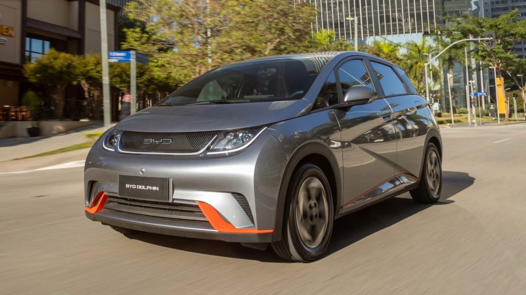 Carro hatchback prata com detalhes laranja, em movimento em uma rua urbana ensolarada.