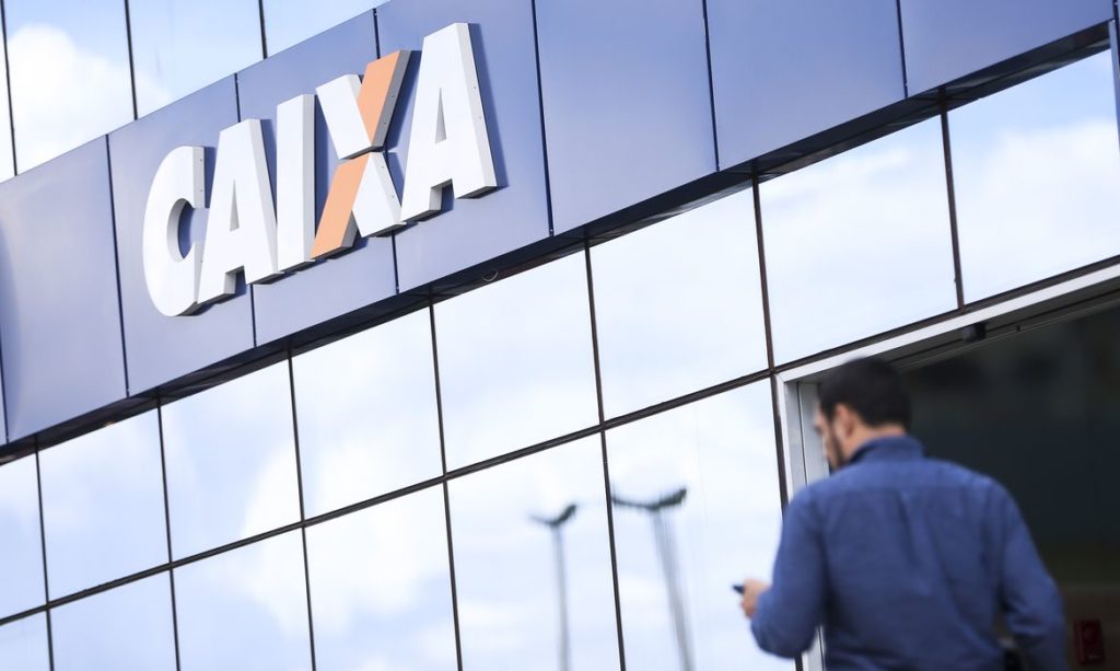 Homem de costas se aproxima da entrada de uma agência da Caixa. O logo "CAIXA" está visível na fachada.