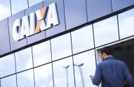 Homem de costas se aproxima da entrada de uma agência da Caixa. O logo "CAIXA" está visível na fachada.