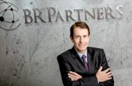 Executivo sorrindo, braços cruzados, em frente à logo BR Partners na parede.