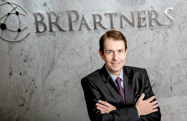 Executivo sorrindo, braços cruzados, em frente à logo BR Partners na parede.