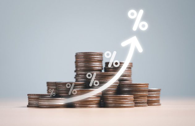 Pilhas de moedas com seta ascendente e símbolos de porcentagem, representando aumento de juros.