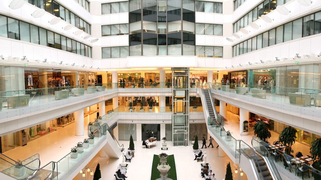 Átrio interno de shopping ou hotel com múltiplos andares, lojas, escadas rolantes, elevadores e pessoas circulando.