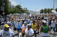 Multidão, majoritariamente mulheres, com camisetas brancas e símbolo do infinito, marchando em rua sob sol.