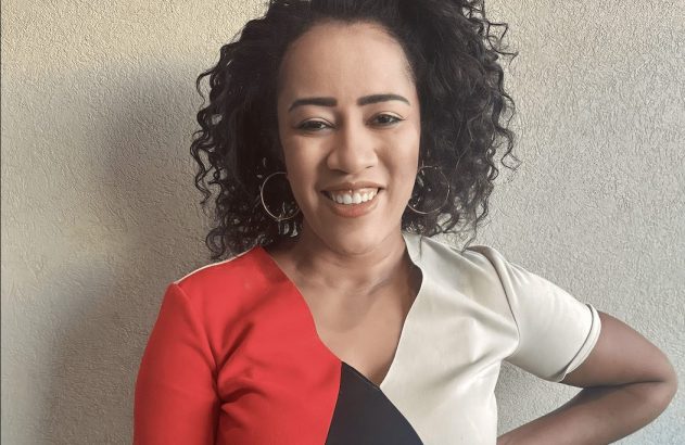 Mulher de cabelo cacheado sorrindo para a câmera, com blusa de bloco de cores e mão no quadril.