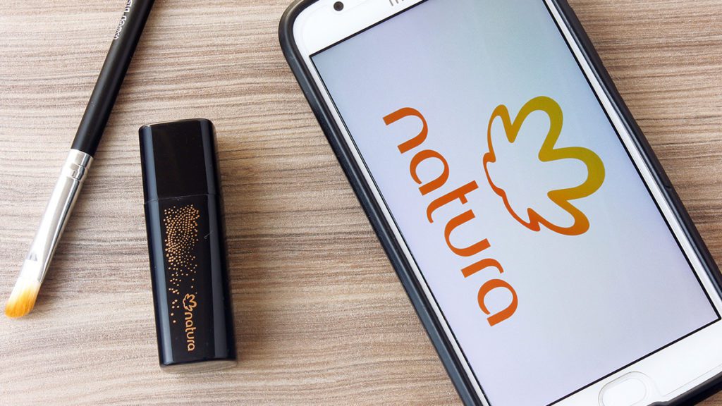 Smartphone com logo Natura na tela, pincel de maquiagem e cosmético preto em mesa de madeira.