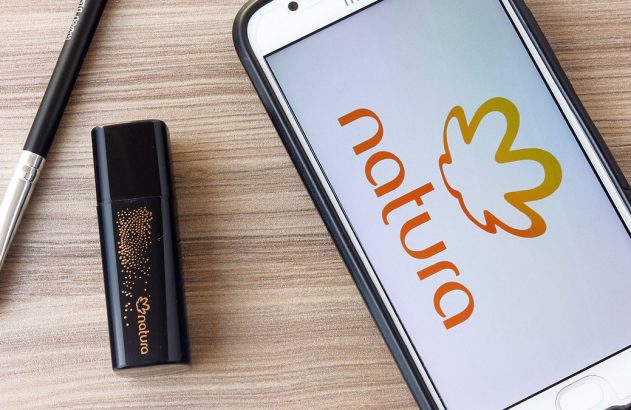 Smartphone com logo Natura na tela, pincel de maquiagem e cosmético preto em mesa de madeira.
