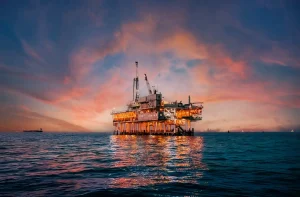 Imagem de uma plataforma de petróleo no mar ao pôr do sol, com céu colorido de tons de rosa e laranja, refletindo na água.
