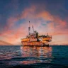Imagem de uma plataforma de petróleo no mar ao pôr do sol, com céu colorido de tons de rosa e laranja, refletindo na água.