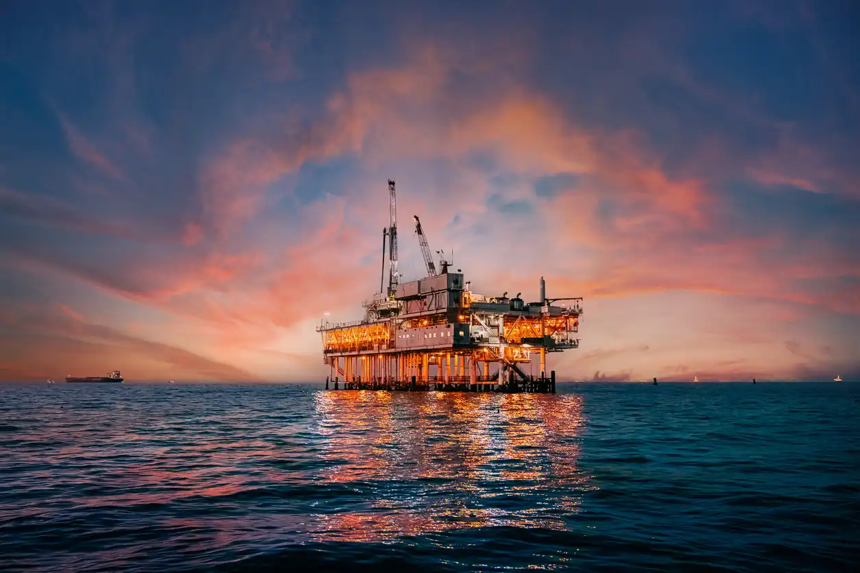 Imagem de uma plataforma de petróleo no mar ao pôr do sol, com céu colorido de tons de rosa e laranja, refletindo na água.
