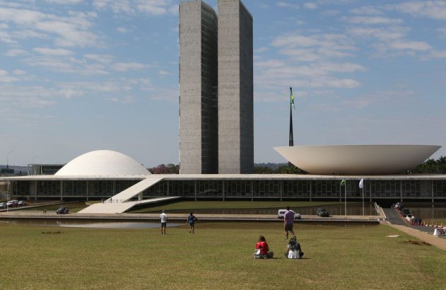O Congresso Nacional do Brasil: edifício branco com duas torres, uma cúpula e uma semiesfera. Pessoas no gramado.