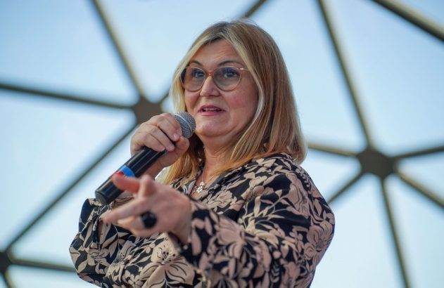 Mulher de óculos fala ao microfone, gesticulando, com fundo de vidro.