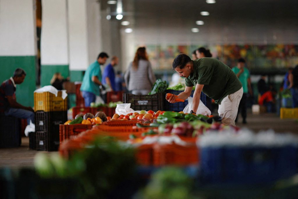 Homem seleciona produtos frescos de caixas em mercado interior, com outras pessoas comprando ao fundo.