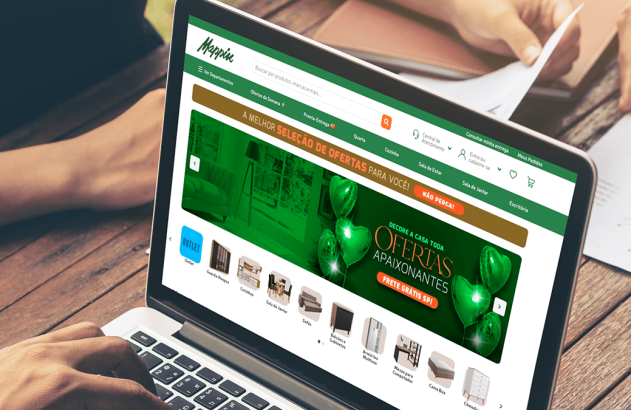 Pessoa digita em laptop exibindo o site Mappin de móveis e ofertas. Mãos de outra pessoa revisam documentos.
