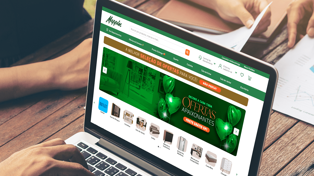 Pessoa digita em laptop exibindo o site Mappin de móveis e ofertas. Mãos de outra pessoa revisam documentos.