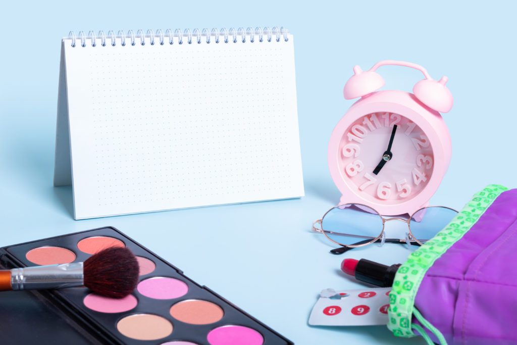 Calendário, despertador rosa e maquiagem para rotina diária ou planejamento, em fundo azul claro.
