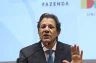 Homem de terno falando ao microfone, com a mão levantada. Ao fundo, painel com "FAZENDA" e "UNIÃO".