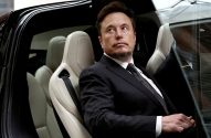 Elon Musk sentado no banco traseiro de um carro, de terno, olhando pensativo para a direita.