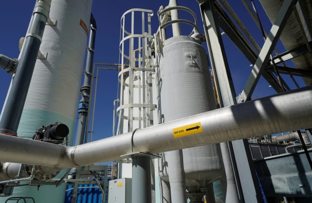 Vista de uma instalação industrial, com grandes tanques e tubulações. Uma etiqueta 'BIO-GAS' aponta o fluxo.