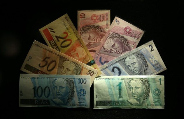 Várias notas de Real brasileiro (1, 2, 5, 10, 20, 50, 100) organizadas em semi-círculo sobre fundo escuro.