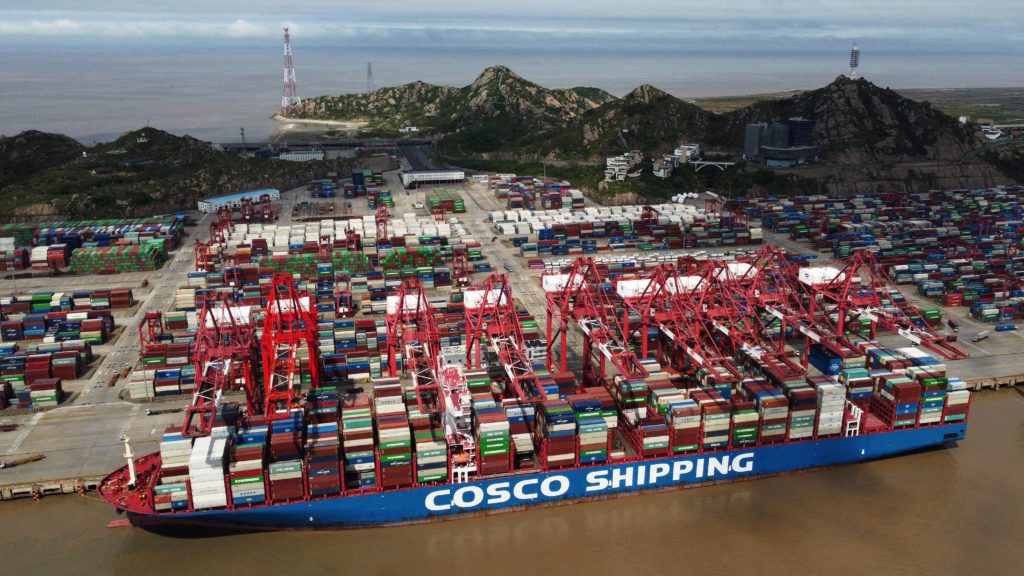 Navio COSCO SHIPPING em porto, guindastes movimentam contêineres. Vasto pátio de carga e montanhas ao fundo.