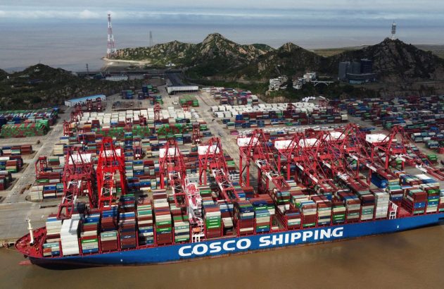 Navio COSCO SHIPPING em porto, guindastes movimentam contêineres. Vasto pátio de carga e montanhas ao fundo.