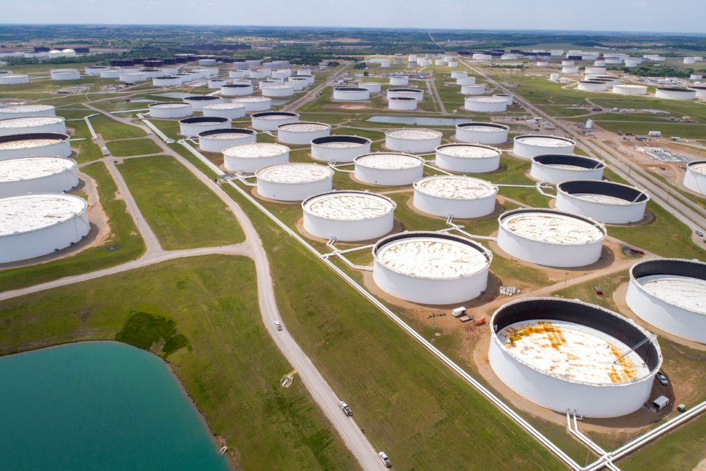 Vista aérea: centenas de grandes tanques de armazenamento de petróleo brancos em um vasto campo verde, com estradas e um lago.