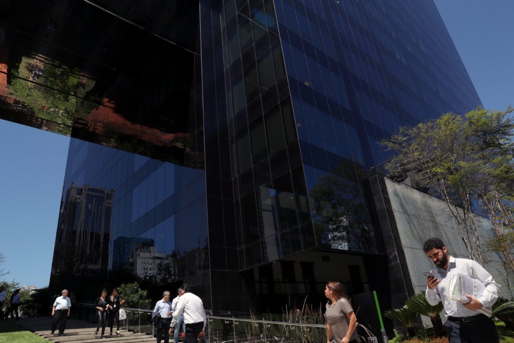 Edifício moderno de vidro escuro reflete céu e árvores. Pessoas caminham nas proximidades sob o sol.