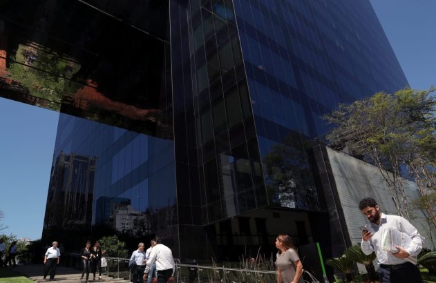 Edifício moderno de vidro escuro reflete céu e árvores. Pessoas caminham nas proximidades sob o sol.