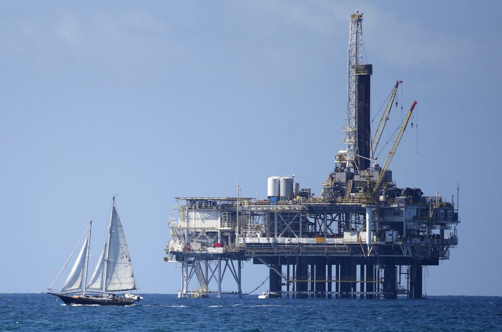 Veleiro navega no mar ao lado de uma plataforma de petróleo offshore, sob um céu azul.