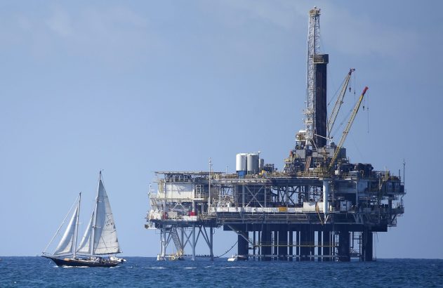 Veleiro navega no mar ao lado de uma plataforma de petróleo offshore, sob um céu azul.